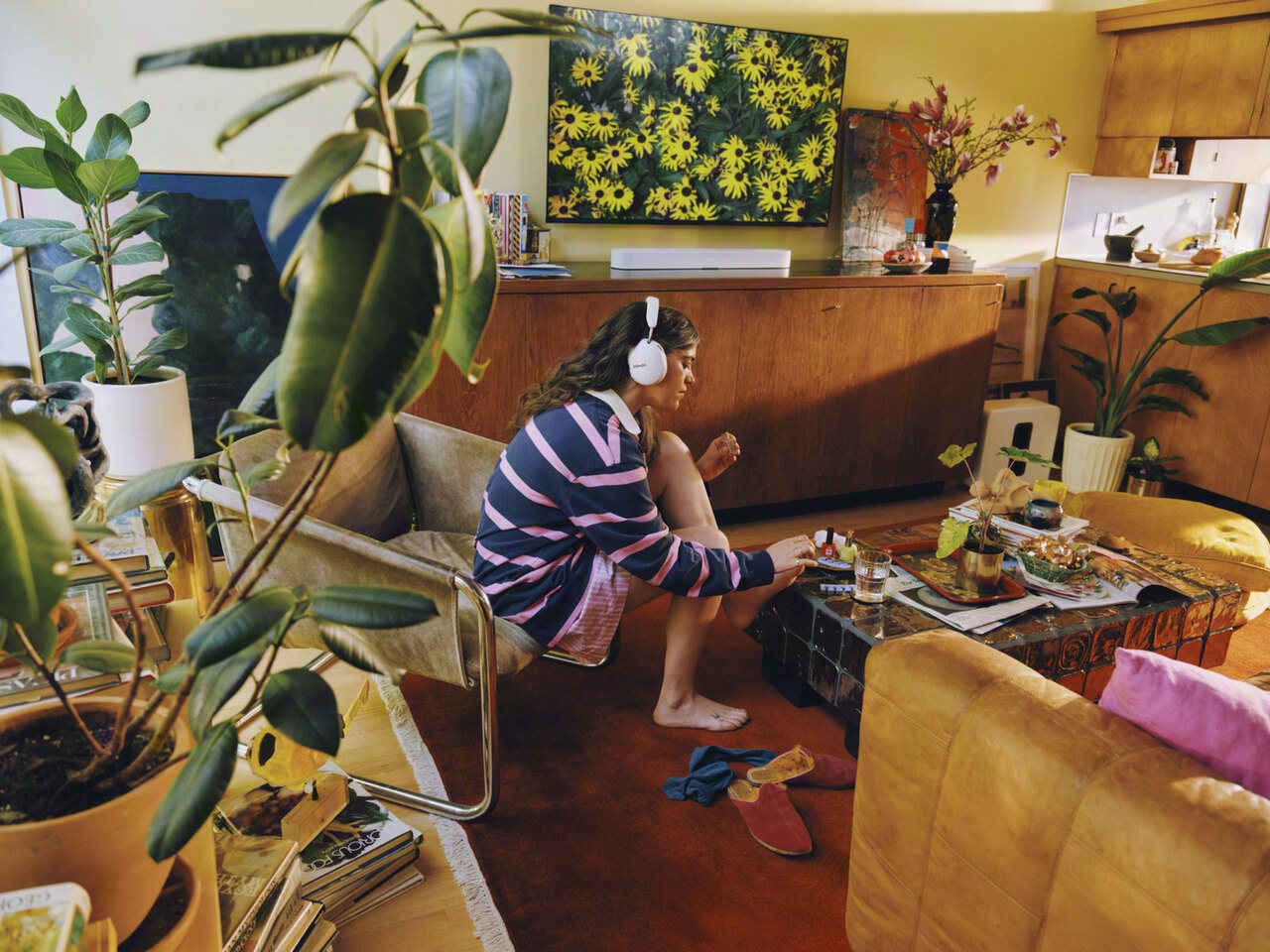 Sonos lifestyle scene with a person wearing white Sonos Ace headphones and a Sonos Beam Ultra soundbar beneath a wall-mounted TV